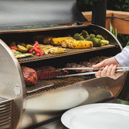 The RT-1250 with the lid open showing a full grill of pork butts, ribeyes, and vegetables with a person using tongs to pull out a pork butt. 